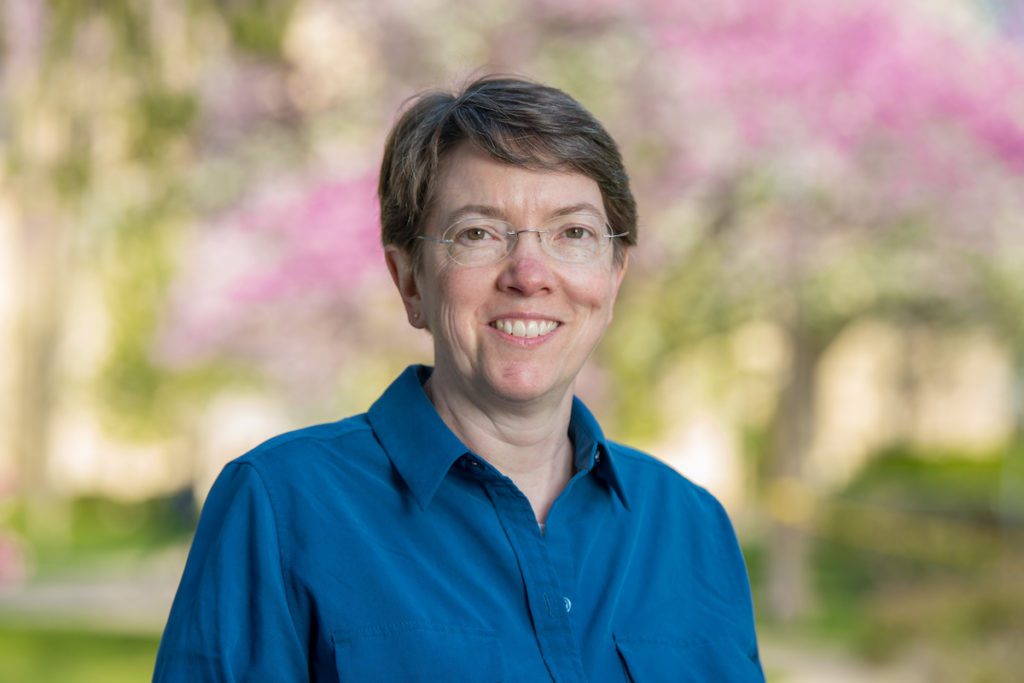 Holly Goodson, a white woman in glasses with short hair and wearing a blue blouse smiles. The background, though out of focus, is the University of Notre Dame campus in spring when trees are in bloom.