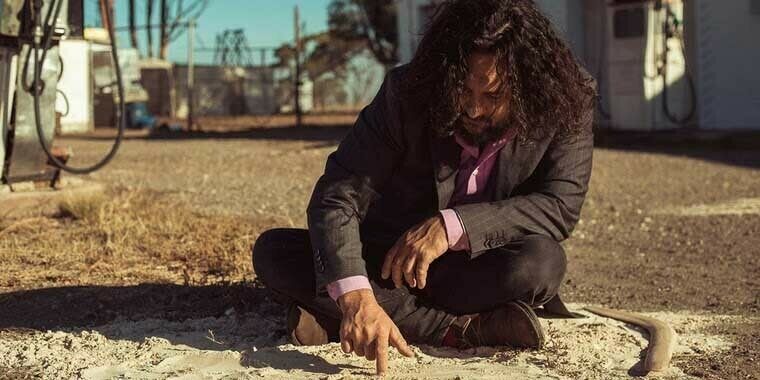 Tyson Yunkaporta, a man with long curly brown hair, and an Australian indigenous author, sits in the sand and demonstrates "Sand Talk."